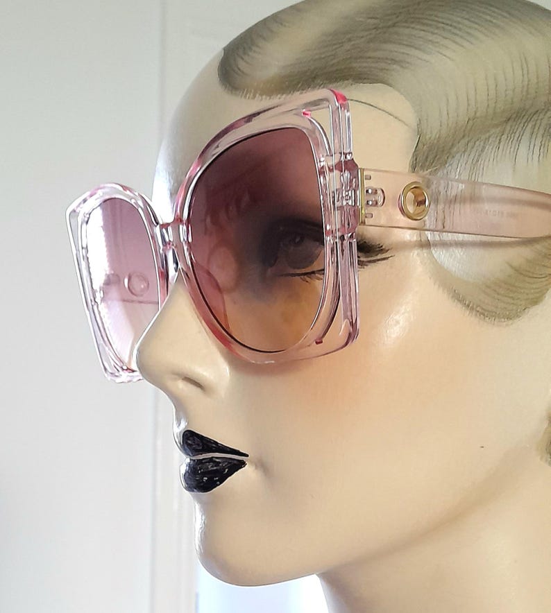 May include: Oversized pink sunglasses with a square frame and gradient lenses. The sunglasses have a light pink frame and temples with a gold-colored accent near the hinges. The lenses transition from a darker pink at the top to a lighter shade at the bottom.