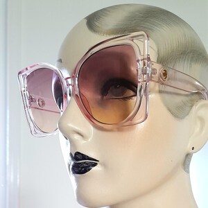 May include: Oversized, square-framed sunglasses in a translucent pink hue. The lenses transition from a light pink to a warm orange. The sunglasses are displayed on a mannequin head, showcasing their bold, retro design.