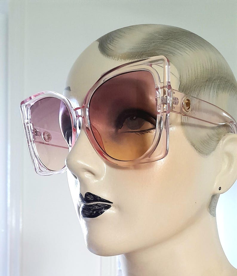 May include: Oversized, square-framed sunglasses in a translucent pink hue. The lenses transition from a light pink to a warm orange. The sunglasses are displayed on a mannequin head, showcasing their bold, retro design.