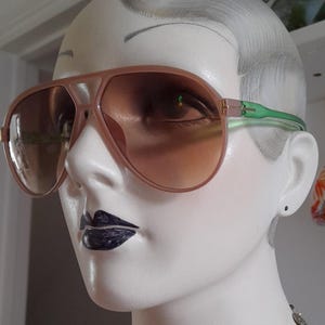 May include: A pair of pink aviator sunglasses with brown lenses and a green frame. The sunglasses are on a mannequin's head.