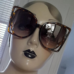 May include: Large, square-framed sunglasses with a tortoiseshell pattern. The sunglasses have a unique, open-frame design around the lenses. The lenses are gradient, transitioning from a darker shade at the top to a lighter shade at the bottom.
