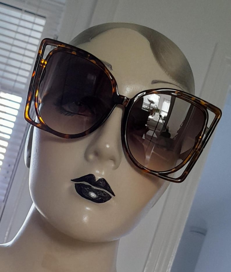 May include: Large, square-framed sunglasses with a tortoiseshell pattern. The sunglasses have a unique, open-frame design around the lenses. The lenses are gradient, transitioning from a darker shade at the top to a lighter shade at the bottom.