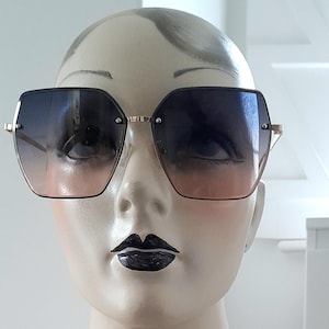 Puede incluir: Gafas de sol geométricas de gran tamaño con una lente degradada que pasa del azul al rosa. Las monturas son doradas y las gafas de sol tienen un diseño moderno y elegante. Las gafas de sol se muestran en una cabeza de maniquí.
