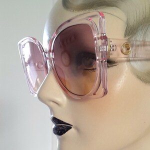 May include: Pink oversized sunglasses with a square frame and gradient lenses. The sunglasses are displayed on a mannequin head, showcasing the design and style. The frames have a transparent appearance, and the lenses transition from a darker shade at the top to a lighter hue.