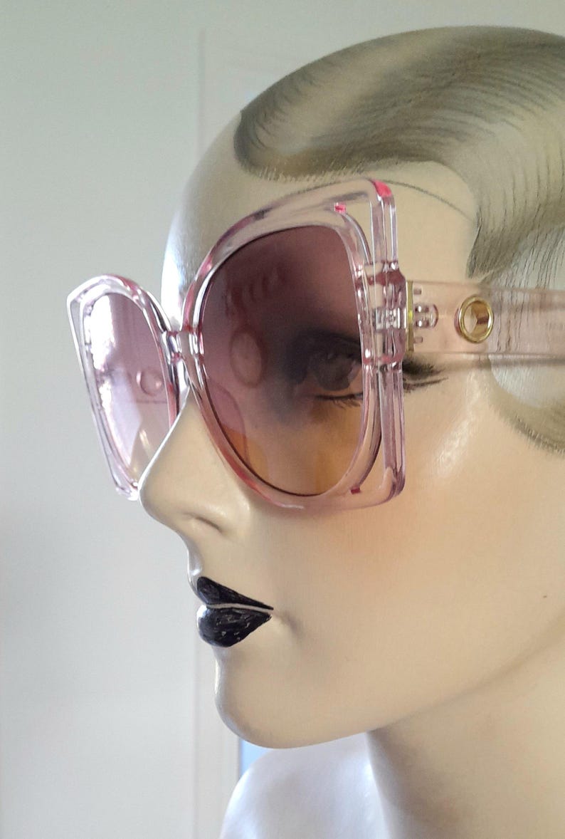 May include: Pink oversized sunglasses with a square frame and gradient lenses. The sunglasses are displayed on a mannequin head, showcasing the design and style. The frames have a transparent appearance, and the lenses transition from a darker shade at the top to a lighter hue.