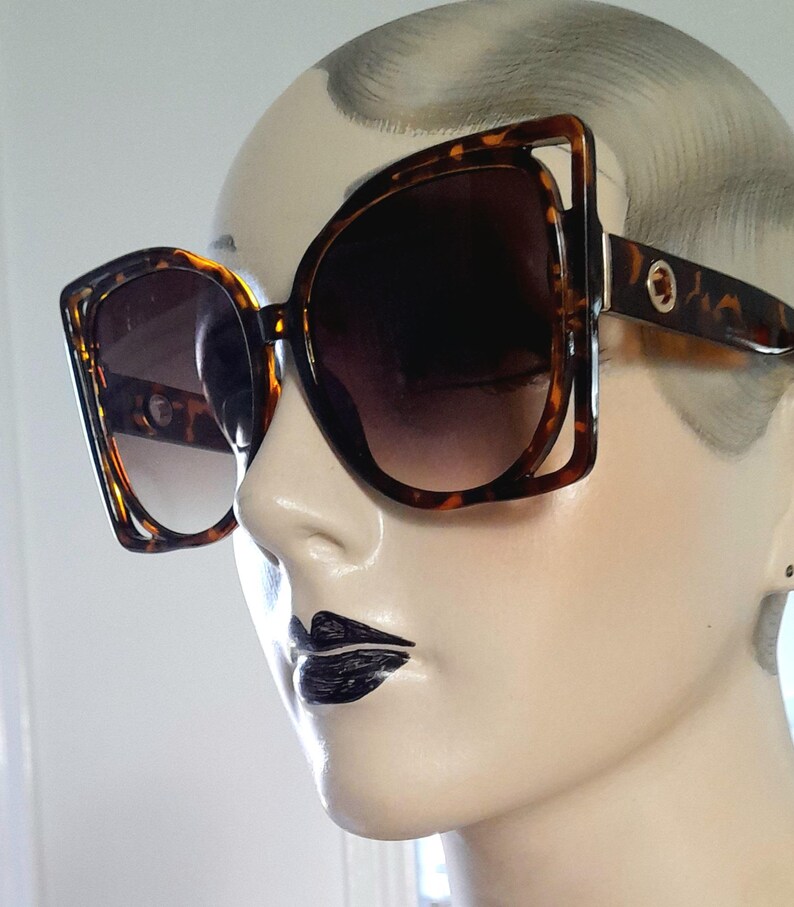 May include: Large, square-framed sunglasses with a tortoiseshell pattern. The lenses are dark, providing shade. The sunglasses are on a mannequin head, showcasing the eyewear's design and style.