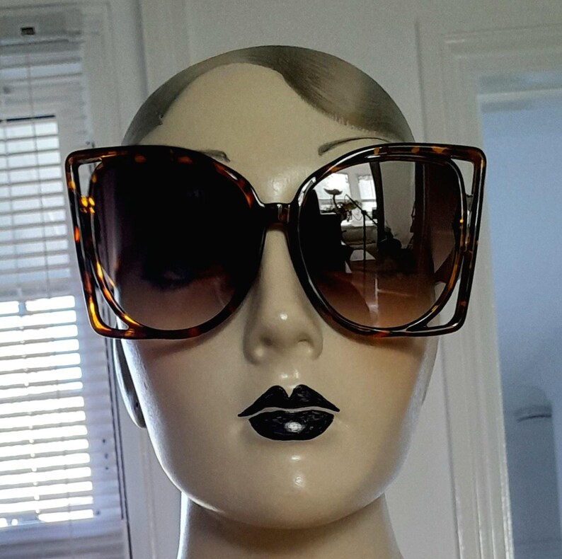 May include: Large, square-framed sunglasses with a tortoiseshell pattern. The lenses are a gradient brown, and the frames have a unique cutout design. The sunglasses are displayed on a mannequin head.