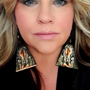 Pode incluir: Brincos triangulares com um design de faraó egípcio. Os brincos têm um esquema de cores dourado e preto com uma ilustração detalhada do rosto de um faraó. Estão pendurados num fundo preto.