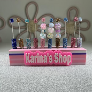 May include: A collection of decorative, bedazzled skewers in various colors, including blue, gold, pink, and teal. Each skewer has a decorative top and sits in a matching bedazzled base. The base is pink and has the text "Karina's Shop".