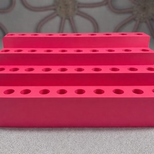May include: A tiered, bright pink display stand with multiple circular holes. The stand has three levels, each with a row of evenly spaced holes. The stand is made of a solid material and is designed to hold small items.