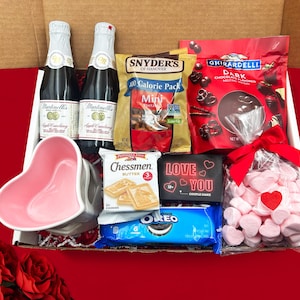 May include: A Valentine's Day gift box filled with treats, including two bottles of sparkling apple cider, a bag of mini pretzels, a bag of dark chocolate melting wafers, a heart-shaped ceramic bowl, a box of Chessmen butter cookies, a couple's game called "Love You", a package of Oreo cookies, and a bag of pink heart-shaped marshmallows.