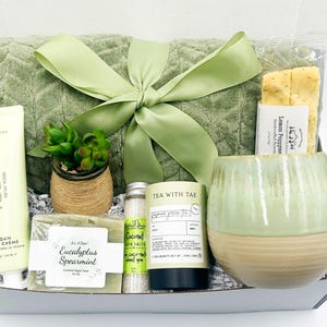 May include: A green and white gift basket with a green towel, a mug, a bar of soap, a jar of bath salts, a bottle of tea, a small potted succulent, and a package of lemon poppyseed tea. The basket is tied with a green ribbon.