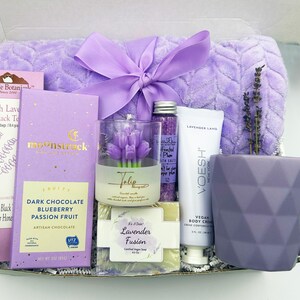 May include: A lavender-themed gift box filled with various items, including a purple mug, a bar of soap, a candle, bath salts, body cream, and a chocolate bar. The box is tied with a lavender ribbon and includes a lavender-colored throw blanket.
