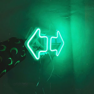 Pode incluir: Um letreiro de luz verde neon em forma de duas flechas apontando em direções opostas. O letreiro está pendurado em uma parede verde.