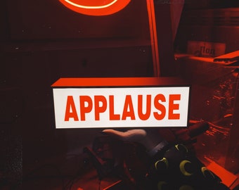 May include: A rectangular light box sign with a white face and a black frame, featuring the word APPLAUSE printed in bold red capital letters. The sign is illuminated, casting a bright red glow in a dark environment with a neon red light above.