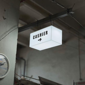 May include: A rectangular, illuminated white sign suspended from a dark metal beam, featuring the black text "CASHIER" above a right-pointing arrow. To the left, a round, white-framed analog wall clock is mounted on a concrete pillar.