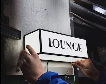 Pode incluir: Uma placa retangular de parede com uma face branca iluminada e uma moldura preta. A palavra LOUNGE está impressa em letras maiúsculas pretas em negrito na superfície frontal. A placa está posicionada horizontalmente contra uma superfície vertical.