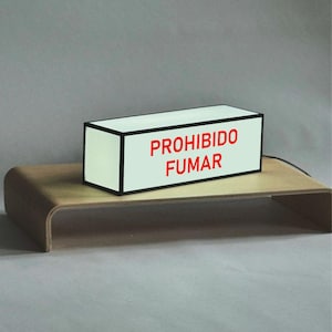 Könnte beinhalten: Ein rechteckiges weißes Schild mit schwarzem Rand und dem roten Text "PROHIBIDO FUMAR". Das Schild steht auf einem geschwungenen Holzständer vor grauem Hintergrund. Das Schild ist wahrscheinlich ein Warnhinweis.