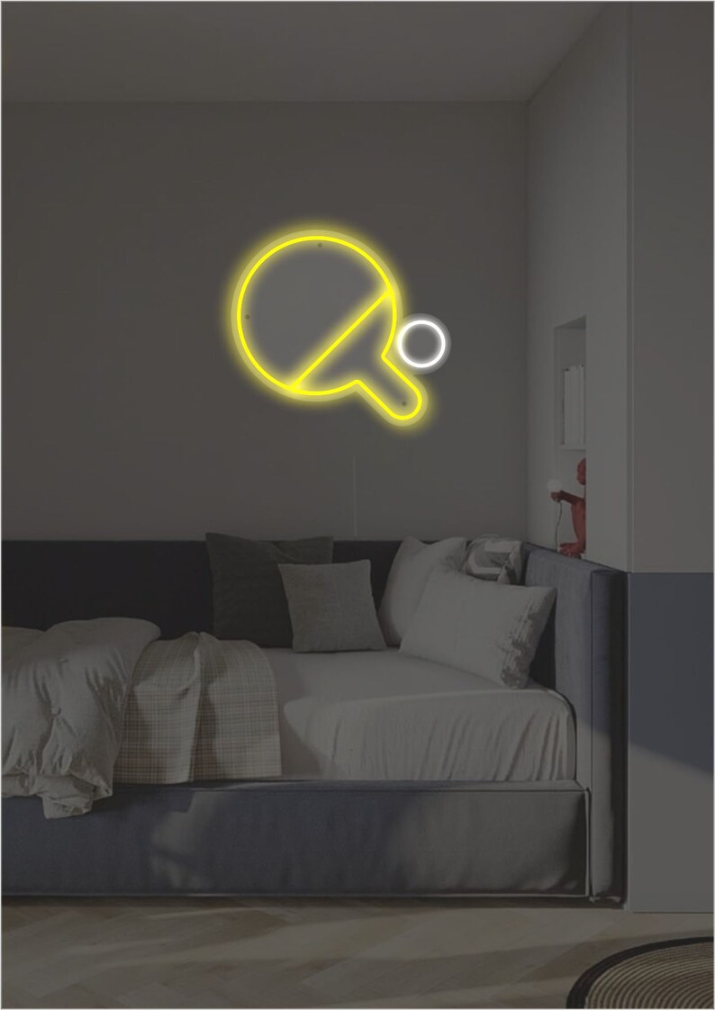 May include: A yellow neon sign of a ping pong paddle and ball on a dark wall above a bed with white bedding.