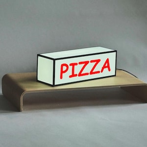 May include: A white box with a black border and the word "PIZZA" in red letters. The box is sitting on a wooden surface.