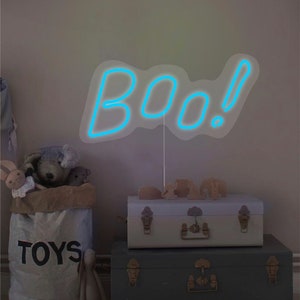 May include: A blue neon sign that spells "BOO!" is the focal point of this image. The sign is propped up in front of a light-colored wall. Below the sign are two decorative trunks, a bag of toys, and stuffed animals. The trunks are stacked on top of each other.