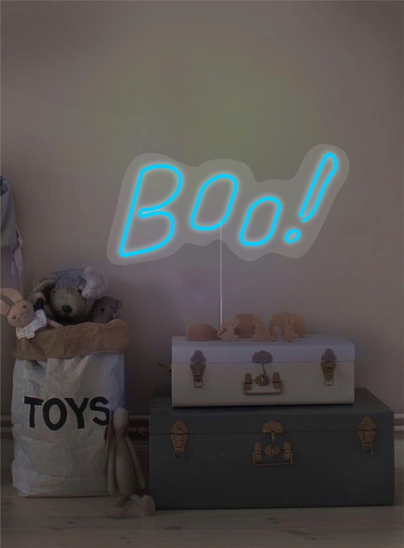 May include: A blue neon sign that spells "BOO!" is the focal point of this image. The sign is propped up in front of a light-colored wall. Below the sign are two decorative trunks, a bag of toys, and stuffed animals. The trunks are stacked on top of each other.