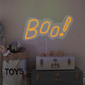 May include: A neon sign in the shape of the word "BOO!" in orange, with a white outline. The sign is placed in a room with a bag of toys, two trunks, and stuffed animals. The bag says "TOYS".