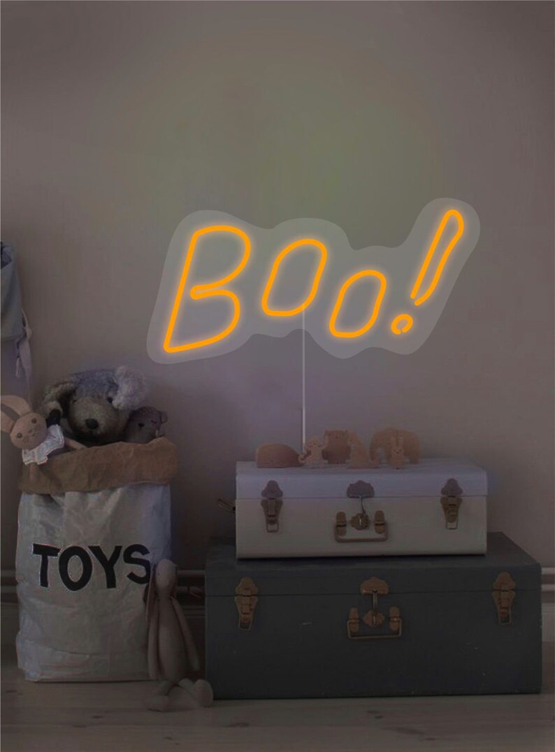 May include: A neon sign in the shape of the word "BOO!" in orange, with a white outline. The sign is placed in a room with a bag of toys, two trunks, and stuffed animals. The bag says "TOYS".