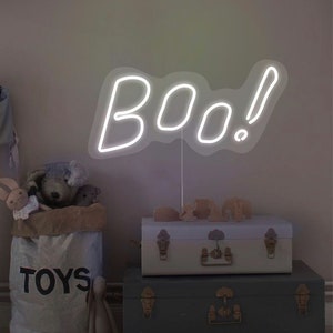 May include: A white neon sign spelling "boo!" stands above two decorative trunks. A canvas bag labeled "TOYS" is filled with stuffed animals. A neutral-toned room with a Halloween theme.