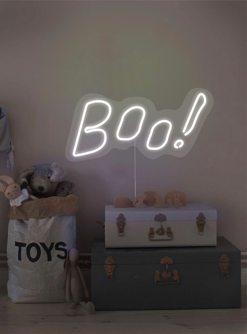 May include: A white neon sign spelling "boo!" stands above two decorative trunks. A canvas bag labeled "TOYS" is filled with stuffed animals. A neutral-toned room with a Halloween theme.