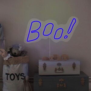 May include: A blue neon "BOO!" sign with an exclamation point, mounted on a white stand. The sign is placed on top of two decorative trunks. A bag of toys and stuffed animals are in the foreground.