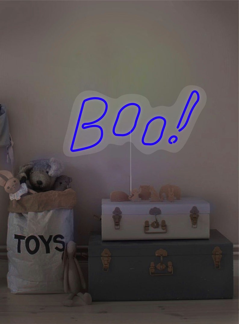 May include: A blue neon "BOO!" sign with an exclamation point, mounted on a white stand. The sign is placed on top of two decorative trunks. A bag of toys and stuffed animals are in the foreground.