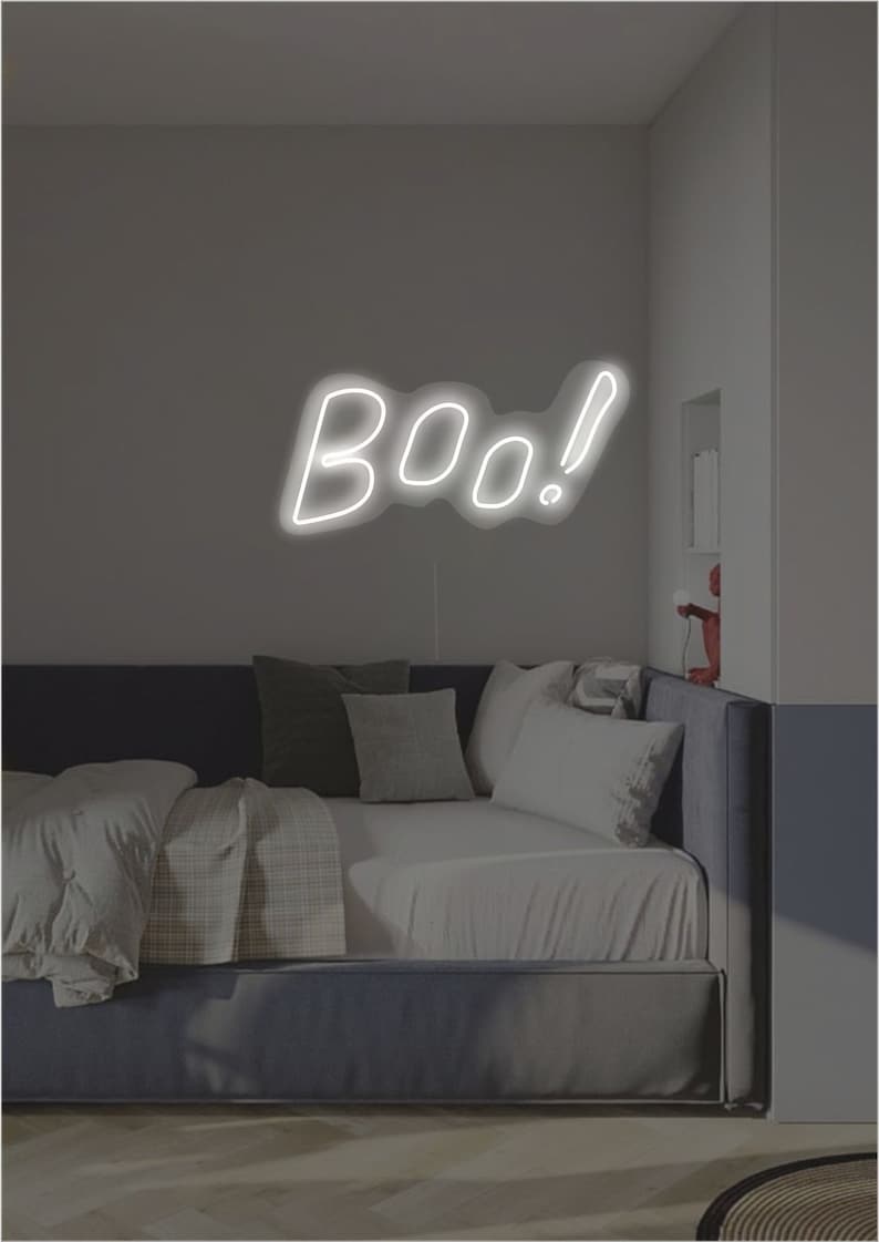May include: A white neon sign in the shape of the word "Boo!" is mounted on a gray wall above a bed with pillows and blankets. The room has a minimalist design with a dark blue bed frame.