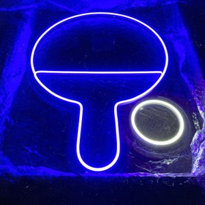 May include: A white neon sign shaped like a ping pong paddle with a white circle next to it. The sign is lit up with a blue glow.
