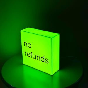 May include: A glowing, neon green rectangular sign with the words "no refunds" in black. The sign is on a dark green table, with a dark green background. The sign is lit from within.