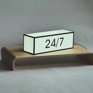 May include: A white light box with a black border and the text "24/7" on the front. The light box is sitting on a wooden shelf.