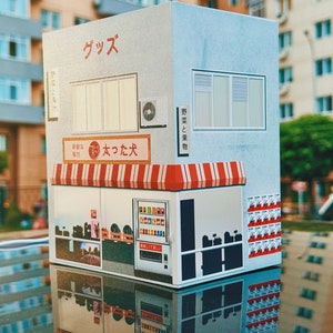 Puede incluir: Un modelo en miniatura de una tienda japonesa con un toldo a rayas rojas y blancas. La tienda tiene una máquina expendedora con una variedad de bebidas y aperitivos. El texto en la tienda dice "グッス" y "大つた大".