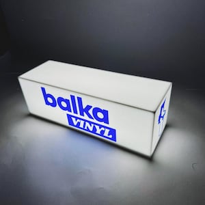 May include: A rectangular, white, illuminated light box sign featuring the blue text "balka" in a bold, lowercase font, with the word "VINYL" printed in white inside a blue rectangular block directly underneath the main text.