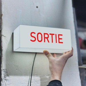 May include: A white rectangular sign with the word "SORTIE" in red letters. The sign is mounted on a textured, off-white wall. A black wire extends from the sign. The sign is being held by a person.