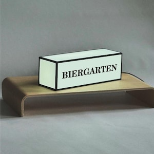 May include: A rectangular light box with the word "BIERGARTEN" in black lettering. The box has a black border and sits on a curved wooden stand. The background is a solid gray.