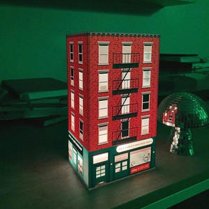 Könnte beinhalten: Ein rotes Backsteingebäude mit einem grünen Vordach und einem Schild mit der Aufschrift "Tony's Famous Original Pizza". Das Gebäude hat fünf Stockwerke und eine Feuertreppe. Das Gebäude ist von innen beleuchtet und im Hintergrund befindet sich eine Discokugel.