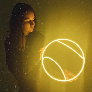 May include: A woman holds a glowing neon yellow tennis ball sign in front of a yellow textured background.