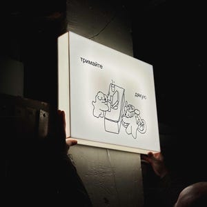 Puede incluir: Un letrero rectangular e iluminado con una ilustración de dibujos animados en blanco y negro. La imagen muestra dos figuras interactuando con una máquina, con texto en un idioma extranjero. El letrero está sostenido por dos manos en un ambiente oscuro.