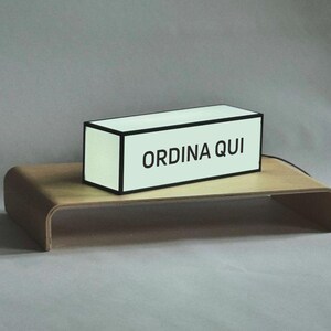 Op de afbeelding: Een rechthoekige lichtbak met de tekst "ORDINA QUI" in zwarte letters, geplaatst op een gebogen houten standaard. De lichtbak heeft een zwarte rand en een lichtgekleurd interieur. De standaard is gemaakt van lichtgekleurd hout.