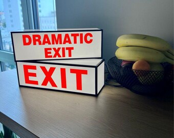 May include: Two rectangular light boxes with black frames and white faces. The top box features the red text "DRAMATIC EXIT" and the bottom box features the red text "EXIT". The boxes are stacked on top of each other on a wooden surface.