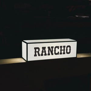 May include: A rectangular, illuminated sign with the word "RANCHO" in bold, black letters. The sign has a white face and a black border, set against a dark background. The sign is lit from within.