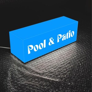 May include: A rectangular, bright blue illuminated sign featuring the white text Pool & Patio. The sign has a clean, box-like structure and emits a soft glow from its base, casting light onto the surface below. A thin white power cord extends from the side.