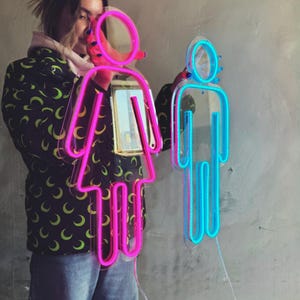 May include: A person holding up two neon signs. The sign on the left is pink and shaped like a person. The sign on the right is blue and shaped like a person.