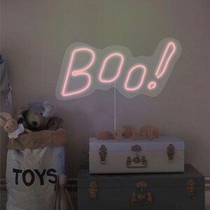 May include: A pink neon sign that spells "boo!" is the focal point of this image. The sign is mounted on a thin white stand. The sign is placed above two decorative trunks and a bag of toys.
