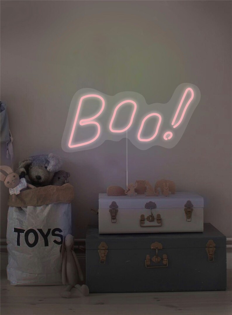 May include: A pink neon sign that spells "boo!" is the focal point of this image. The sign is mounted on a thin white stand. The sign is placed above two decorative trunks and a bag of toys.